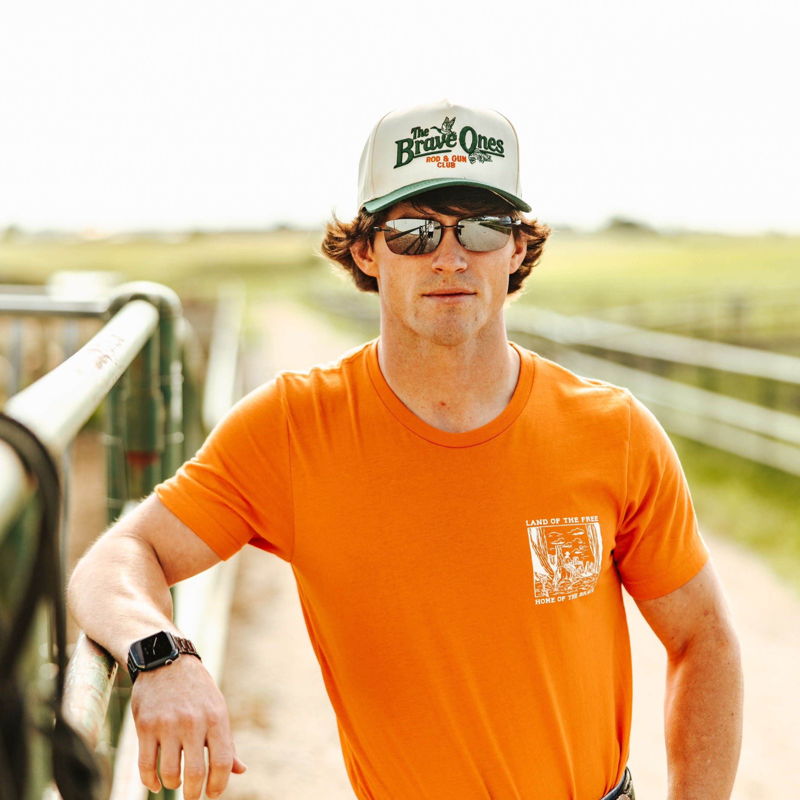 Man wearing an orange shirt and sunglasses, standing on a dirt path with a scenic background.