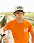Man wearing an orange shirt and sunglasses, standing on a dirt path with a scenic background.
